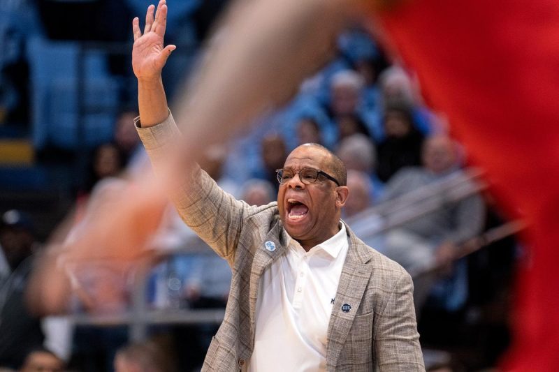 Hubert Davis signals to his players while raising his hand. He's wearing a gray, plaid pinstripe dress jacket over a white shirt with his glasses on.